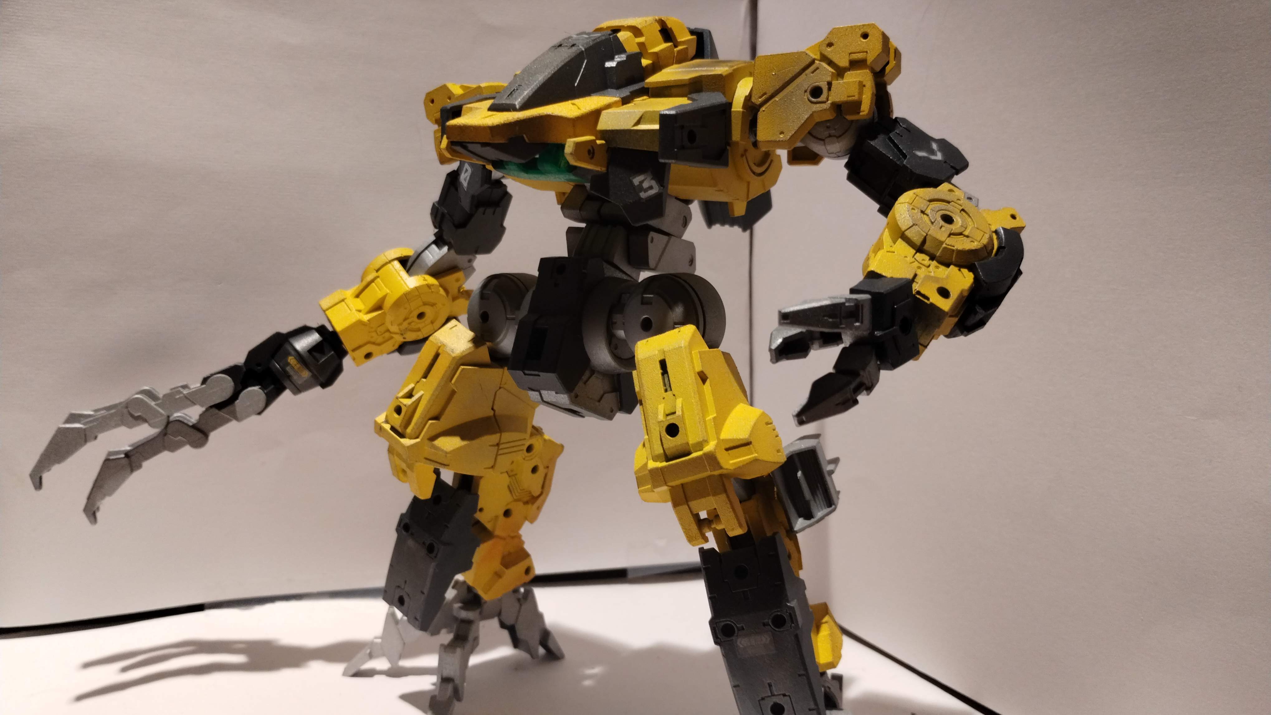 A painted model kit of a bulky robot with no face. The unit is matte yellow in the color of common construction equipment with black accents. The bodywork is covered in warning stickers, gunsmoke, dust and dirt.